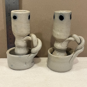 May include: Two handmade ceramic candle holders. Each has a cylindrical body with three dark blue dots and a handle. The candle holders are light gray with a textured surface.