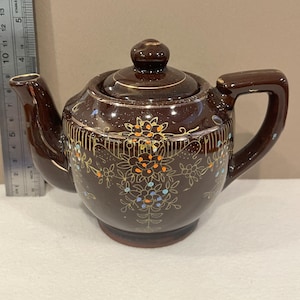 May include: A brown ceramic teapot with a lid and handle. The teapot is decorated with floral patterns in orange, blue, and gold. The teapot is approximately 6 inches tall.