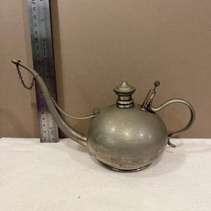 May include: A silver-colored metal oil lamp with a rounded body, a curved spout, and a handle. The lamp has a small chain attached to the spout and a lid on top. The lamp is approximately 6 inches long.