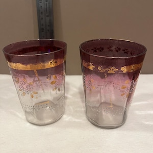 May include: Two vintage drinking glasses. The glasses have a clear base that fades to pink and then to a deep burgundy rim. A gold band and floral design adorn the upper portion of the glasses. The glasses are likely made of glass.
