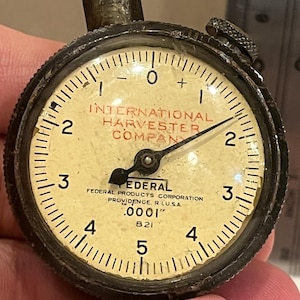 May include: Close-up of a vintage dial indicator with a black bezel and a cream-colored face. The face features the text "INTERNATIONAL HARVESTER COMPANY" and "FEDERAL". The dial measures in increments of .0001 inches.
