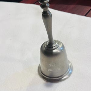 May include: A silver-colored bell with a rounded base and a handle. The bell has the text "TASC 1966 1986" engraved on its side. The handle has a decorative, tiered design at the top.