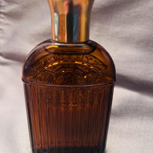 Puede incluir: Un frasco de perfume de vidrio marr&oacute;n con una tapa dorada. La botella tiene un cuerpo redondeado con texto en relieve y una base acanalada y c&oacute;nica. La tapa es de color dorado brillante. La botella es probablemente vintage o antigua.