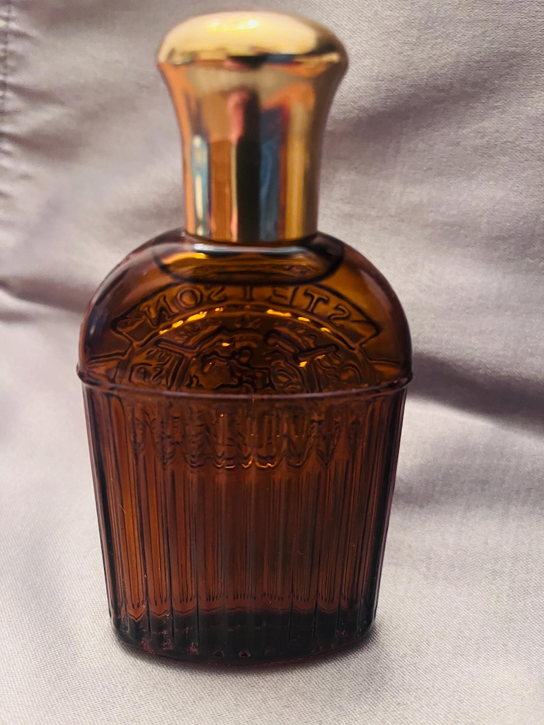 Puede incluir: Un frasco de perfume de vidrio marr&oacute;n con una tapa dorada. La botella tiene un cuerpo redondeado con texto en relieve y una base acanalada y c&oacute;nica. La tapa es de color dorado brillante. La botella es probablemente vintage o antigua.