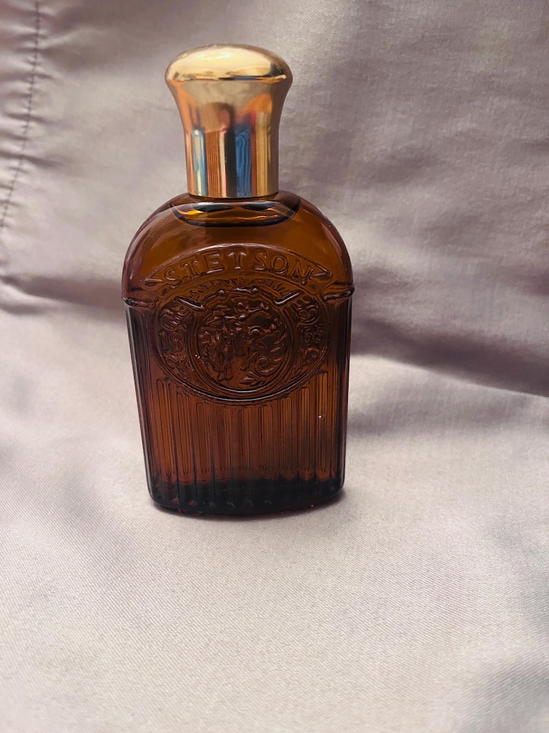 Puede incluir: Una botella de vidrio marr&oacute;n con una tapa dorada. La botella tiene un dise&ntilde;o texturizado y la palabra "STETSON" est&aacute; grabada en relieve en la parte delantera. Probablemente un frasco de perfume o colonia.