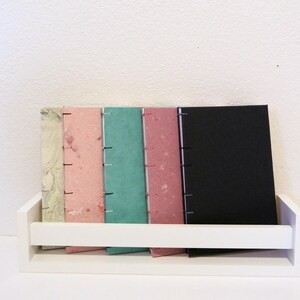 May include: A white shelf displays a collection of handmade journals. The journals have covers in various colors, including marbled, pink, teal, and black. Each journal is bound with visible stitching along the spine, creating a rustic aesthetic.