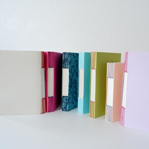 May include: A collection of handmade journals with colorful covers. The journals are bound with visible stitching along the spine. The covers are in various colors, including pink, teal, light blue, green, peach, and lavender. The journals are standing upright.
