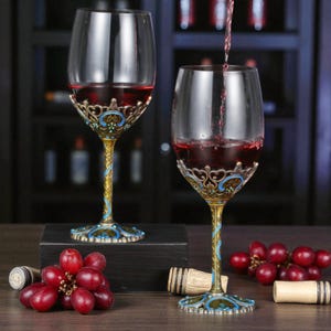 Puede incluir: Dos copas de vino ornamentadas con vino tinto, con tallos dorados y detalles de filigrana azul y dorada. Un racimo de uvas rojas y corchos están sobre la mesa. Una copa se está llenando.