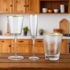 May include: Three clear drinking glasses with a textured surface and gold rims are displayed on a wooden table. The glasses include a wine glass, a champagne flute, and a tumbler. The background shows a kitchen setting.