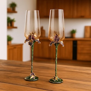 Könnte beinhalten: Zwei Sektgläser mit bernsteinfarbenen Kelchen und aufwendigen floralen Stielmustern. Die Stiele sind grün mit einem gedrehten Muster, und die floralen Akzente sind gold- und lilafarben. Auf einer Holzoberfläche platziert.
