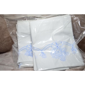 Puede incluir: Sábanas blancas con un diseño floral y de lazo azul, empaquetadas en plástico transparente. Las sábanas están dobladas y parecen nuevas. El diseño está impreso en un delicado color azul.
