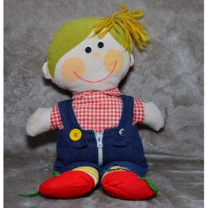 Może przedstawiać: Zabytkowa szmaciana lalka z uśmiechniętą twarzą, czarnymi oczami z guzików i różowymi policzkami. Lalka ma na sobie czerwoną i białą koszulę w kratę, niebieskie dżinsowe ogrodniczki z żółtym guzikiem i czerwone buty. Lalka ma żółte włosy z przędzy.