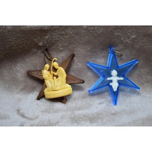 May include: Two Christmas tree decorations. One is a brown wooden star with a cream-coloured nativity scene. The other is a blue star with a white baby Jesus figure inside. Both have metal loops for hanging.