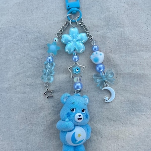 Może przedstawiać: Niebieski brelok do kluczy Care Bear z motywem księżyca i gwiazdy na brzuchu. Brelok posiada niebieskie zapięcie, łańcuszek, koraliki, gwiazdki, kwiat, kokardę i zawieszkę w kształcie półksiężyca. Paleta kolorów to głównie jasnoniebieski i srebrny.