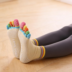 Puede incluir: Calcetines de yoga beige con ranuras individuales para los dedos, cada uno rematado con un color diferente. Los calcetines tienen una suela antideslizante y las palabras "LOVE YOGA" impresas en la suela. Los puños de los calcetines tienen rayas arcoíris.