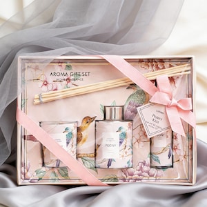 Op de afbeelding: Aroma cadeau set met een kaars, diffuser en geurolie. De set heeft een bloemen- en vogelontwerp met het woord "Peony" op de flessen. Een roze lint is om de doos gebonden.