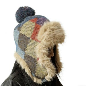 May include: A colorful winter hat with a patchwork design in shades of blue, red, yellow, and gray. The hat features a faux fur trim and a pom-pom on top. The hat is designed to cover the ears.