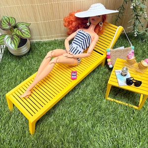 May include: A doll with curly red hair wearing a white and navy blue striped swimsuit and a wide-brimmed white hat, reclining on a yellow slatted lounge chair. A small pink smartphone, a straw bag with colorful pom-poms, and sunglasses are nearby.