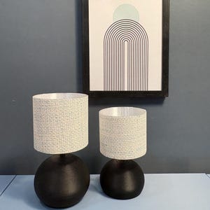 May include: Two small black table lamps with patterned cylindrical shades. A framed print with an abstract design of a semi-circle and arches hangs on a grey wall. The lamps are on a light blue surface.