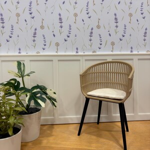 May include: A light brown wicker chair with black legs and a cream-colored cushion. The chair is set against a white wall with a lavender and key pattern wallpaper. Two potted plants are in the foreground.