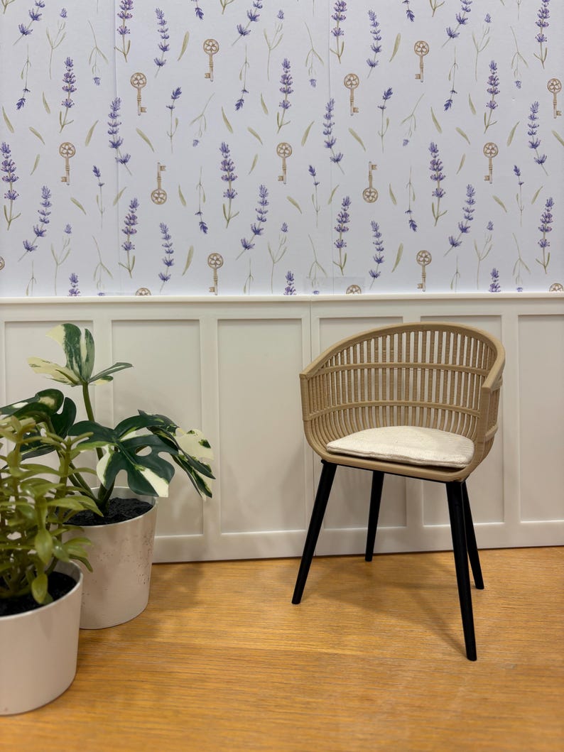 May include: A light brown wicker chair with black legs and a cream-colored cushion. The chair is set against a white wall with a lavender and key pattern wallpaper. Two potted plants are in the foreground.