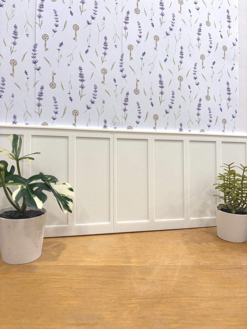 May include: A decorative wall with a white wainscoting and a wallpaper featuring lavender sprigs and antique keys. Two potted plants sit on a wooden surface, adding a touch of greenery to the scene.