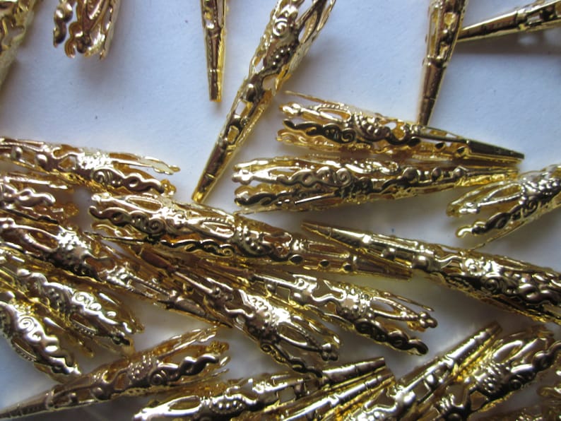 May include: A pile of gold-colored metal tips with intricate designs. The tips are pointed and have a decorative, ornate look.