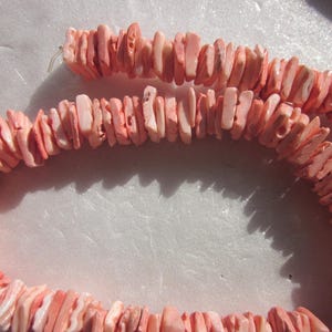 Puede incluir: Una hebra de cuentas de coral rosa, cada cuenta es plana y de forma irregular. Las cuentas están enhebradas para formar un collar o una pulsera.