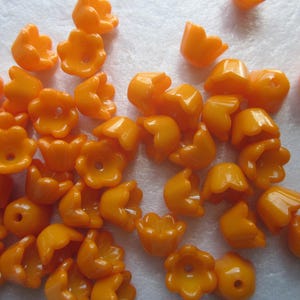 May include: A collection of small, orange, flower-shaped beads. Each bead has a hole in the center and a glossy finish. The beads are scattered across a light-colored surface, showcasing their vibrant color and unique shape.