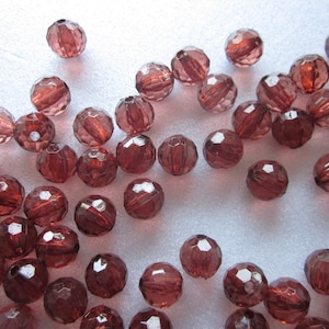 May include: A collection of faceted glass beads in a deep burgundy color. The beads are scattered on a white surface.