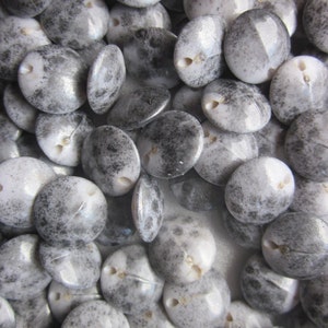 May include: A close-up of a pile of round, white and grey buttons with a speckled pattern. The buttons have two holes each and are made of a shiny, slightly iridescent material.