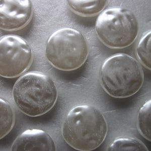 May include: A collection of 13 white, round, pearl-like buttons. The buttons are smooth and have a slightly iridescent sheen.