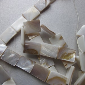 May include: Strand of rectangular, iridescent mother-of-pearl beads. The beads are white with subtle brown and gold hues, strung together with a thin, clear cord. The beads are approximately 1 cm in length, suitable for jewelry making.