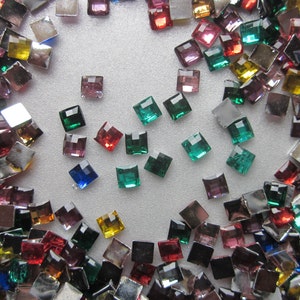 May include: Assortment of colourful square rhinestones in various shades of red, green, blue, purple, and gold. The rhinestones are scattered on a white surface.