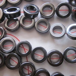 May include: A close-up of a string of black hematite donut beads. The beads are all the same size and shape, and they are strung on a red string.