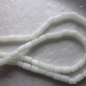 May include: Close-up of a strand of white, rectangular beads. The beads are strung together, forming a necklace or bracelet. The background is a light, textured surface.