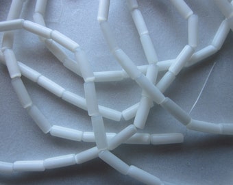 Pode incluir: Close-up de contas cilíndricas brancas enfiadas. As contas são lisas e de tamanho uniforme, criando um padrão delicado e entrelaçado. As contas são provavelmente usadas para fazer joias ou outros projetos de artesanato.