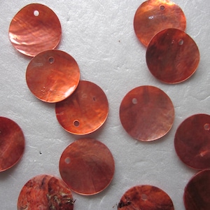 May include: A collection of 12 round, orange-brown, iridescent, mother-of-pearl shell discs with a hole in the center. The discs are approximately 1 inch in diameter.