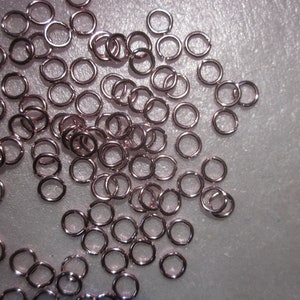 May include: A large quantity of small, rose gold jump rings. The rings are all the same size and are scattered on a white surface.