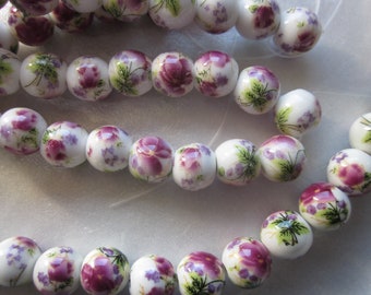 Può includere: Una collana o un braccialetto composto da perline di ceramica bianca con motivi floreali viola e verdi.