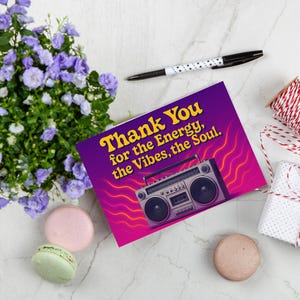 May include: Greeting card with a purple and pink gradient background, featuring a retro boombox illustration and the text "Thank You for the Energy, the Vibes, the Soul." The image also includes a pen, macarons, flowers, and wrapped gifts.