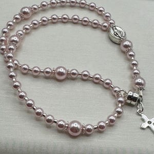 May include: A light pink rosary necklace with pearl beads of varying sizes. The necklace features a silver-toned cross pendant and a circular centerpiece with a religious emblem. The beads are strung together with silver-toned links.