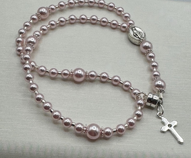 May include: A light pink rosary necklace with pearl beads of varying sizes. The necklace features a silver-toned cross pendant and a circular centerpiece with a religious emblem. The beads are strung together with silver-toned links.