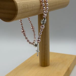 May include: A rosary with pink beads and a silver cross pendant. The rosary is displayed on a wooden stand. The beads are a soft pink color, and the cross is detailed with a small crucifix.