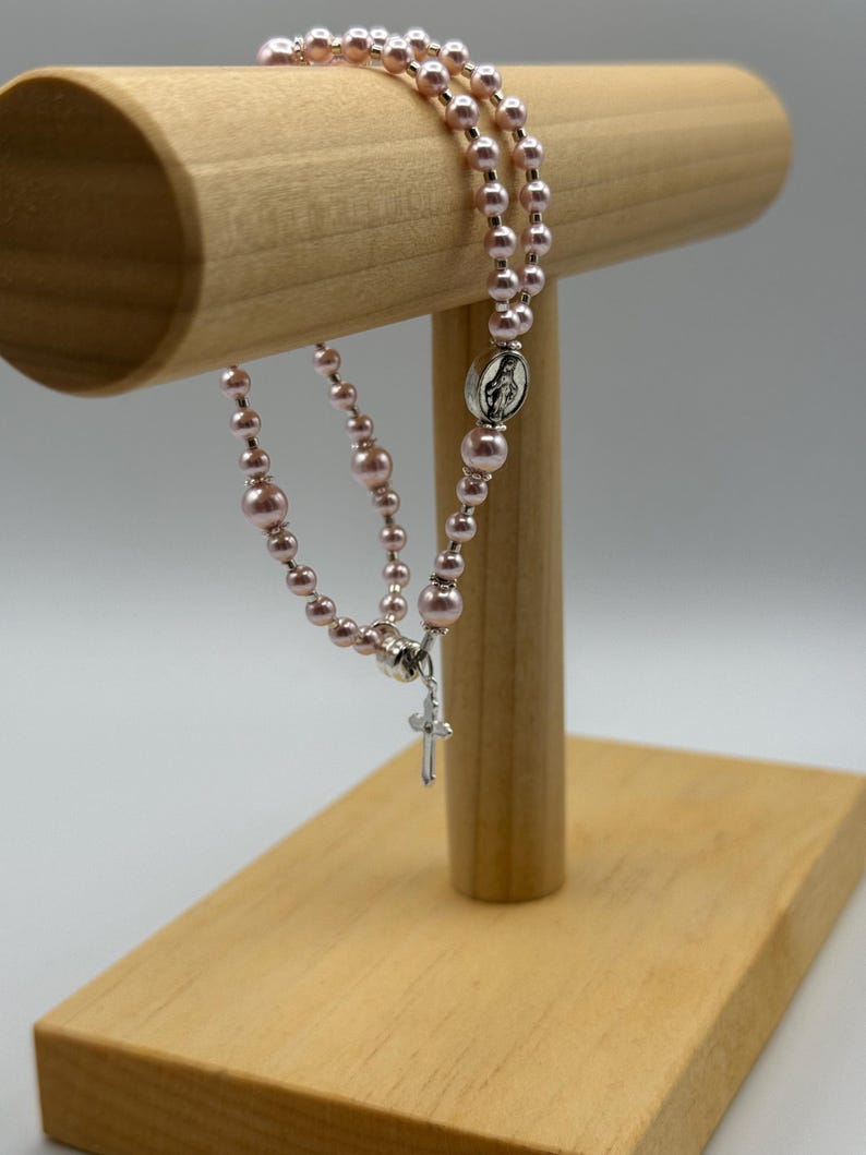 May include: A rosary with pink beads and a silver cross pendant. The rosary is displayed on a wooden stand. The beads are a soft pink color, and the cross is detailed with a small crucifix.