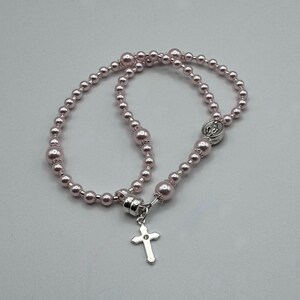 May include: A rosary necklace featuring light pink beads of varying sizes. A silver-colored cross pendant hangs from the clasp. The necklace is designed with a series of beads and a decorative clasp.