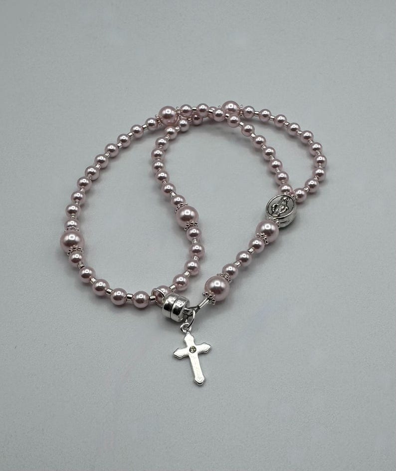 May include: A rosary necklace featuring light pink beads of varying sizes. A silver-colored cross pendant hangs from the clasp. The necklace is designed with a series of beads and a decorative clasp.