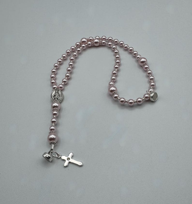May include: A pink beaded rosary with a silver cross pendant and a magnetic clasp. The rosary features a silver centerpiece with a religious figure. The beads are a soft pink color.