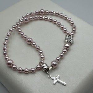 May include: A pink rosary with pearl beads and a silver cross pendant. The rosary features a silver connector and a small medallion with a religious figure. The beads are a soft pink color.