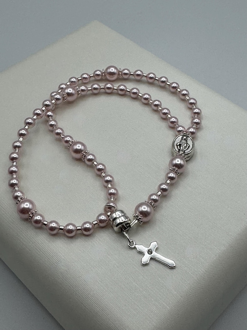 May include: A pink rosary with pearl beads and a silver cross pendant. The rosary features a silver connector and a small medallion with a religious figure. The beads are a soft pink color.